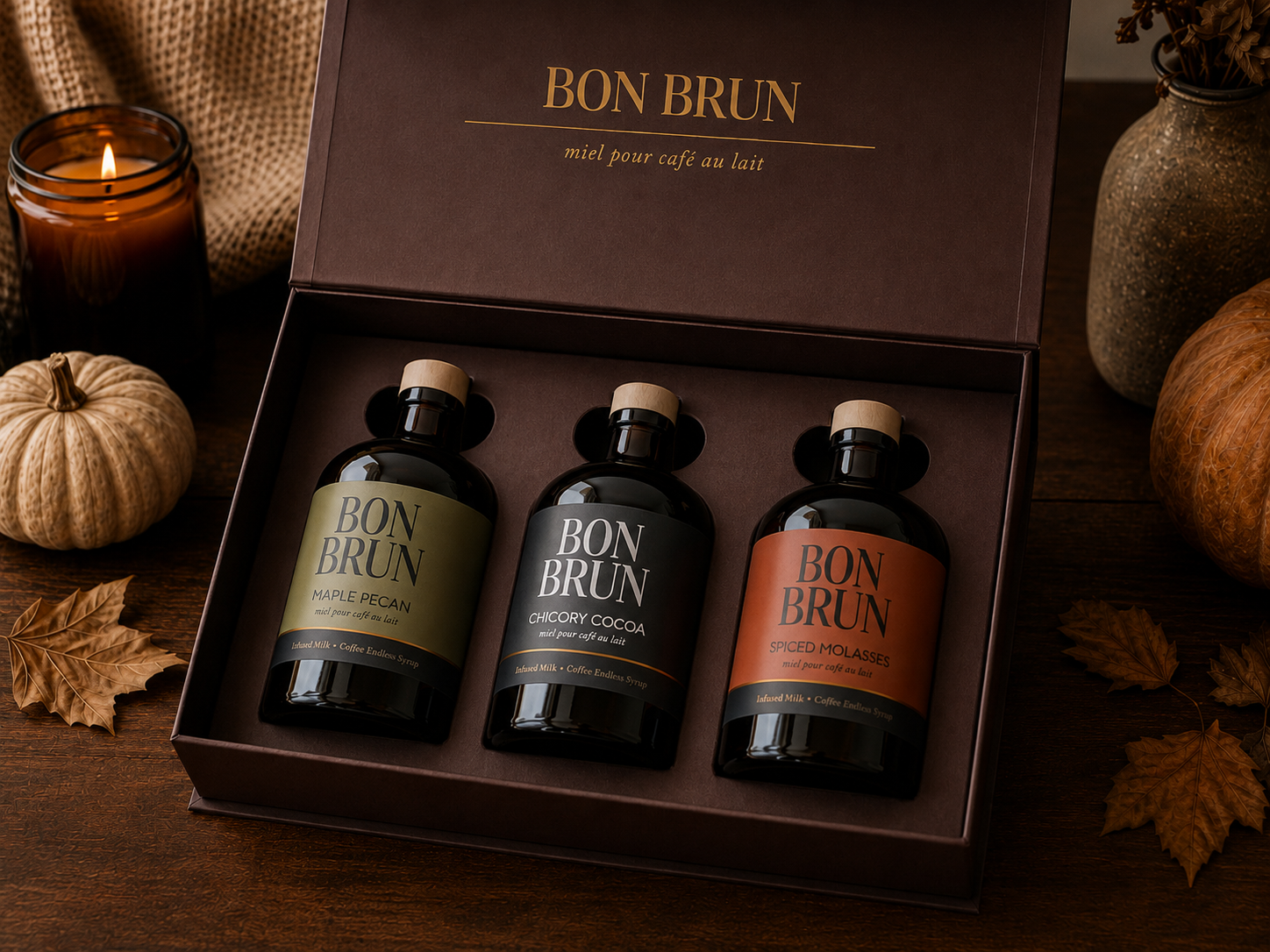 A boxed set of three Bon BruN bottled beverages presented in an open dark brown gift box on a wooden surface, surrounded by pumpkins, leaves, and a lit candle. The bottles have dark glass and labels in olive green, black, and terracotta orange.