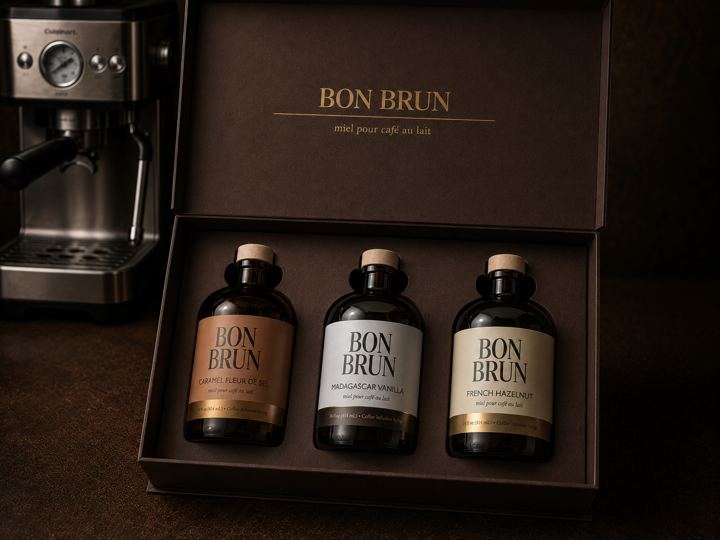 A boxed coffee gift set from BON BRUN, shown open with three dark glass bottles arranged in a dark brown presentation box. A stainless steel espresso machine is partially visible at the upper left.