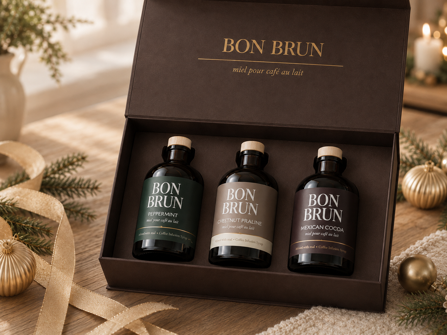 A boxed set of three Bon Brun milk coffee blends presented in a dark brown gift box on a wooden surface with holiday decor. The set includes three dark glass bottles with cream-colored caps, labeled 'BON BRUN' 